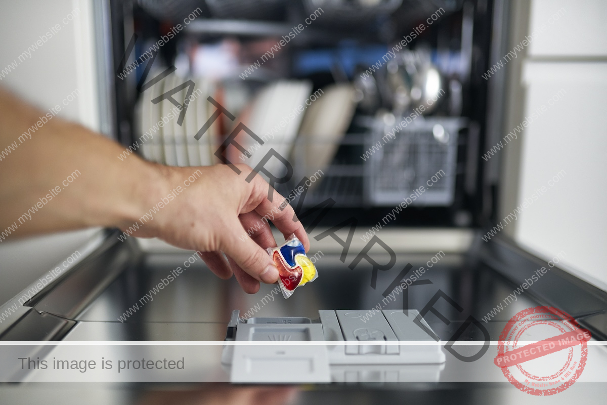 A person's hand putting red, blue and yellow dishwasher pods into an open dishwasher.