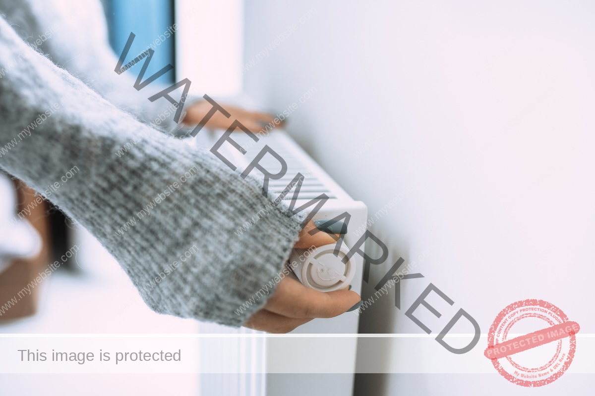 A woman in a gray sweater adjusts an old dial-style thermostat.