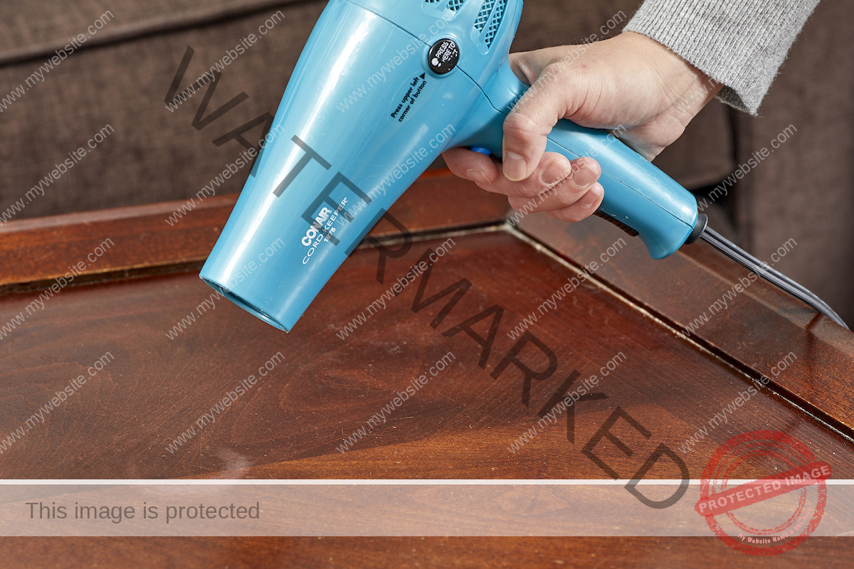 A woman points a blue hair dryer at a heat stain on a wooden coffee table.