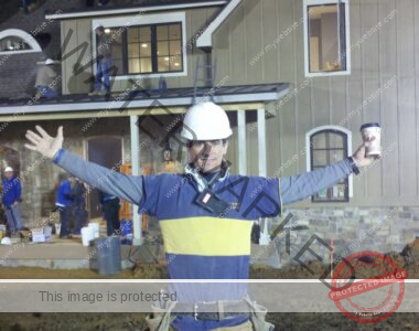 A project manager in a hard hat stands in front of a house being renovated with his arms outstretched.