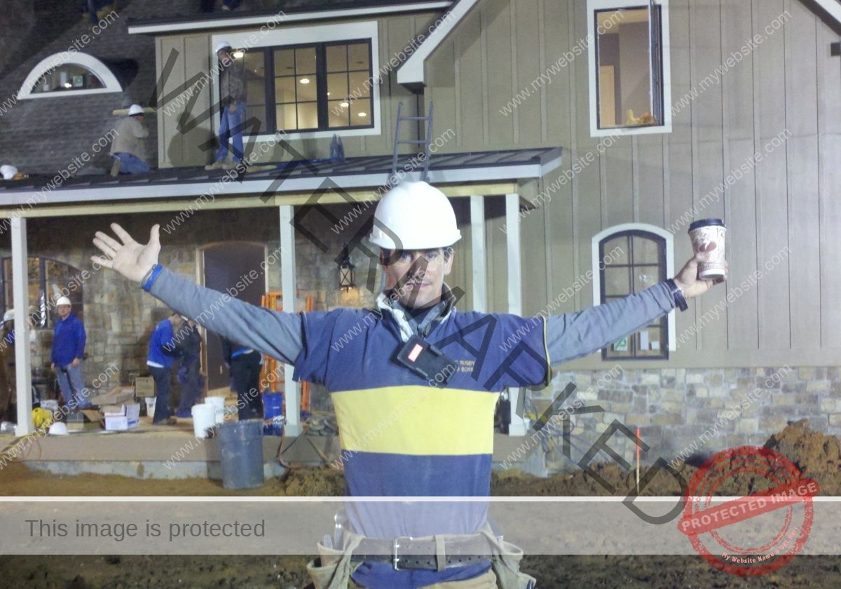 A project manager in a hard hat stands in front of a house being renovated with his arms outstretched.