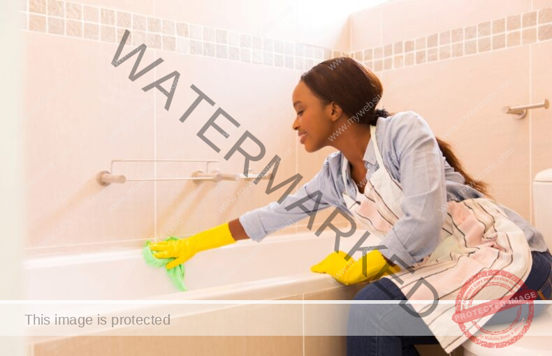 Young woman with her hair tied back cleaning a pink tile surrounded tub.